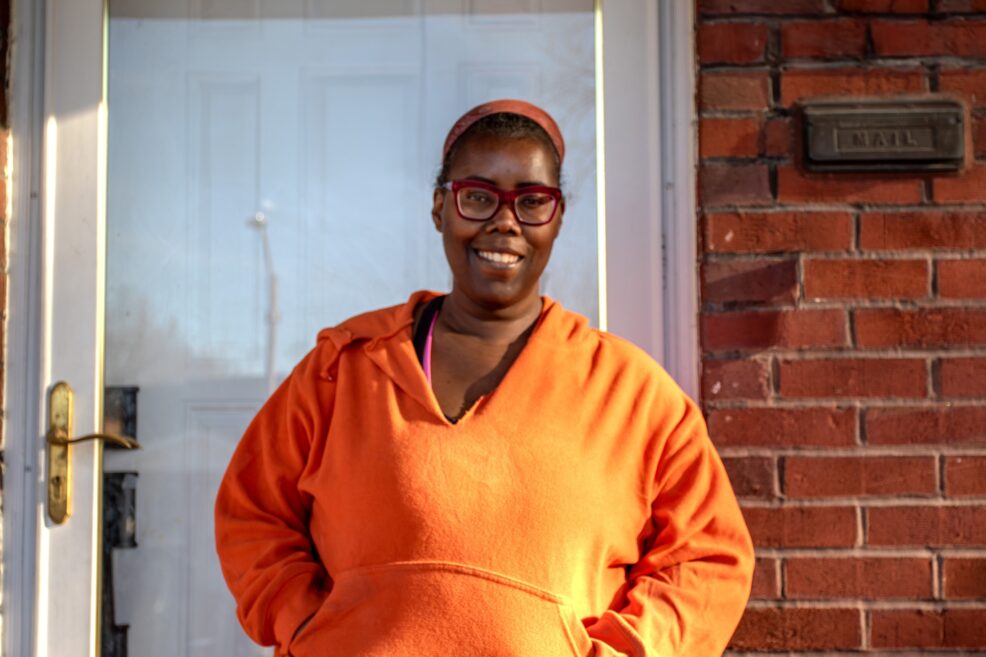 Picture of lady smiling in front of house door wearing red glasses and a orange hoodie.